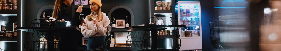 Two women in a stylish caf&eacute;, one with a yellow cap, holding glasses of wine. Shelves with products and a fridge can be seen in the background., &copy; Stuttgart-Marketing GmbH, Alwin Maigler