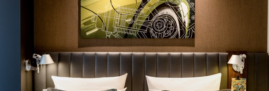 Ein Hotelzimmer mit einem großen Kunstwerk eines Autos über dem Bett. Das Bett hat graue Kissen und eine gepolsterte Rückwand., © TOMAS Ein Hotelzimmer mit einem großen Kunstwerk eines Autos über dem Bett. Das Bett hat graue Kissen und eine gepolsterte Rückwand., © TOMAS