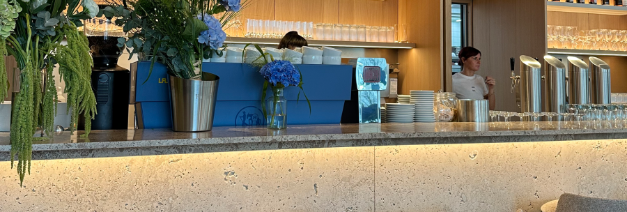 Modern bar with blue and white flower arrangements, drinks on the shelf and gray bar stools. One person stands behind the counter., © Stuttgart-Marketing GmbH