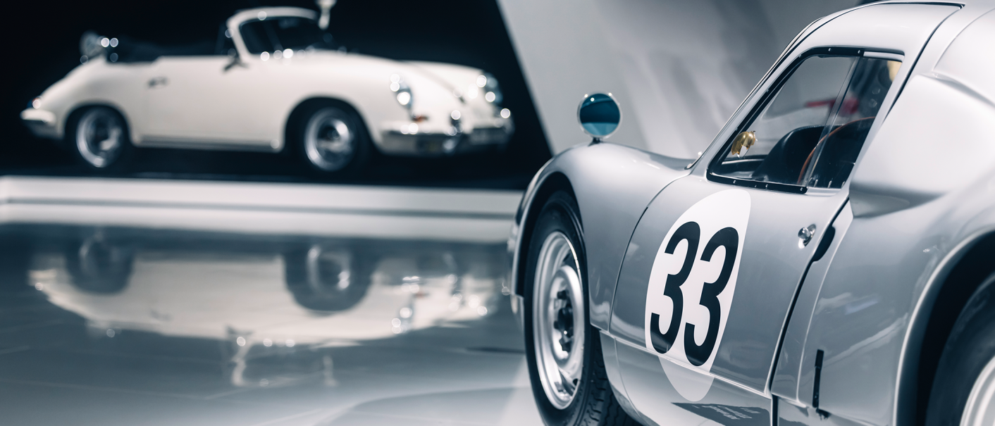 Two classic cars in an exhibition. A white convertible with police lights and a silver car with the number 33 in the foreground., © Porsche AG Two classic cars in an exhibition. A white convertible with police lights and a silver car with the number 33 in the foreground., © Porsche AG