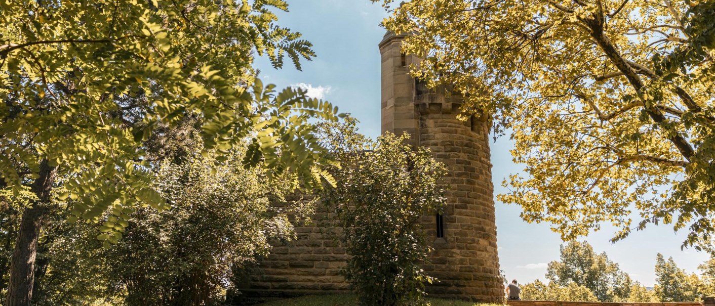 Ein Steinturm, teilweise verdeckt von herbstlichen Bäumen, unter einem klaren blauen Himmel. Eine Person sitzt auf einer Mauer., © Stuttgart Marketing GmbH, Sarah Schmid