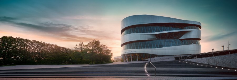 Das Mercedes-Benz Museum in Stuttgart bei Sonnenuntergang. Moderne Architektur mit geschwungenen Linien, umgeben von Bäumen und beleuchteten Stufen., © Mercedes-Benz Heritage GmbH