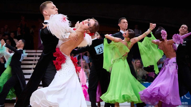 Tanzpaare in eleganten Kost&uuml;men tanzen bei der German Open Championship. Frauen tragen bunte Kleider, M&auml;nner schwarze Anz&uuml;ge. Zuschauer im Hintergrund., &copy; GOC, Fotograf: Bob van Ooik