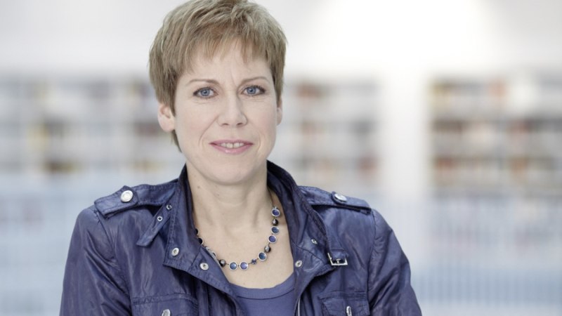 Frau mit kurzem Haar und blauer Jacke vor unscharfem Hintergrund, lächelnd in die Kamera blickend., © Foto: Volker Schrank Frau mit kurzem Haar und blauer Jacke vor unscharfem Hintergrund, lächelnd in die Kamera blickend., © Foto: Volker Schrank
