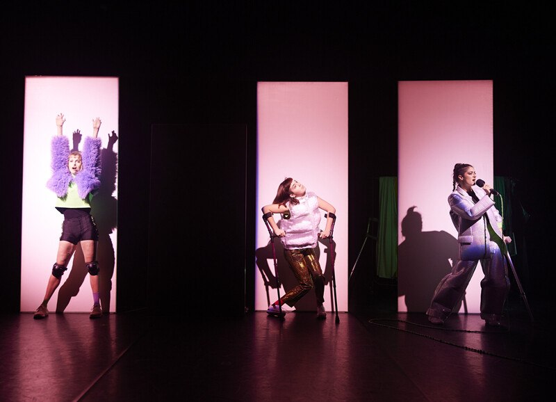 Drei Personen auf einer Bühne in auffälliger Kleidung, beleuchtet von rosa Licht. Eine Person singt in ein Mikrofon, die anderen posieren., © Württembergische Staatstheater Stuttgart