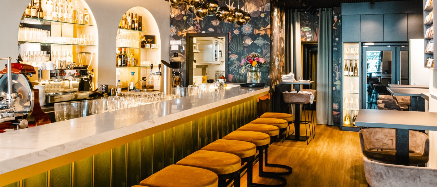 Elegante Bar mit langer Theke, gelben Barhockern und stilvollen Lampen. Die Wände sind mit floralen Mustern dekoriert, Flaschenregale im Hintergrund., © SMG, Sarah Schmid