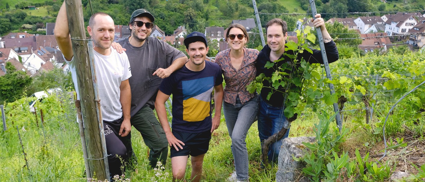 Five people are smiling as they pose in a vineyard. A village can be seen in the background. The group looks relaxed and at one with nature., © Weinkollektiv Edenberg & Tal Five people are smiling as they pose in a vineyard. A village can be seen in the background. The group looks relaxed and at one with nature., © Weinkollektiv Edenberg & Tal