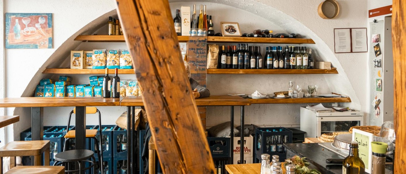 Ein gemütlicher Feinkostladen mit Holzregalen, die Weinflaschen und Pasta präsentieren. Ein Holzbalken dominiert die Mitte des Bildes., © SMG, Sarah Schmid