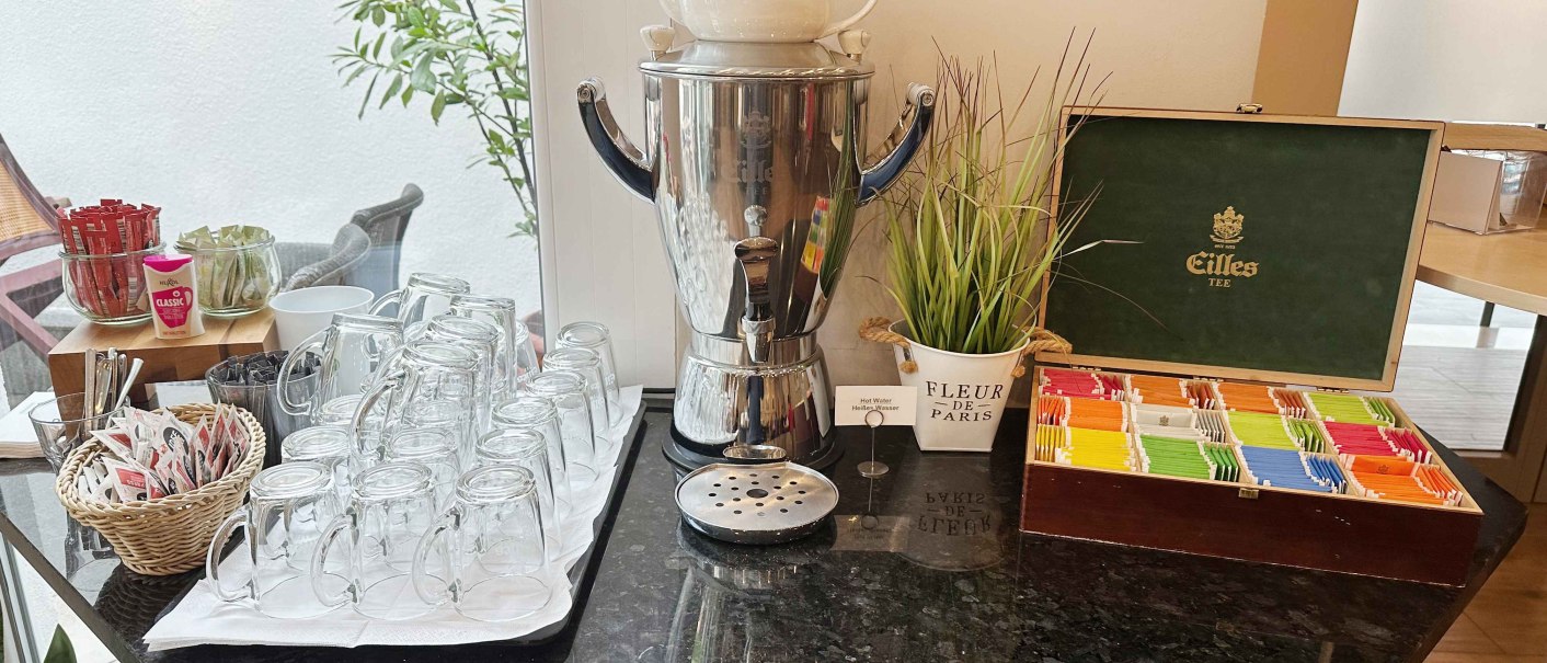 Teestation mit Gl&auml;sern, Teebeuteln und einem Wasserkocher auf einer schwarzen Theke. Dekorative Pflanze und Teebox vorhanden., &copy; Novum Hospitality