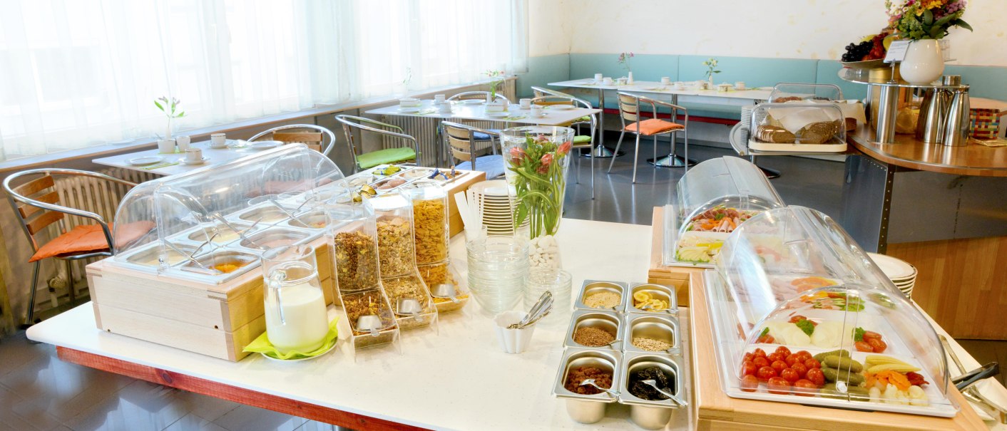 Frühstücksbuffet mit Müsli, Joghurt, Aufschnitt und Obst in einem hellen Raum mit gedeckten Tischen., © Abalon Hotel Ideal Frühstücksbuffet mit Müsli, Joghurt, Aufschnitt und Obst in einem hellen Raum mit gedeckten Tischen., © Abalon Hotel Ideal
