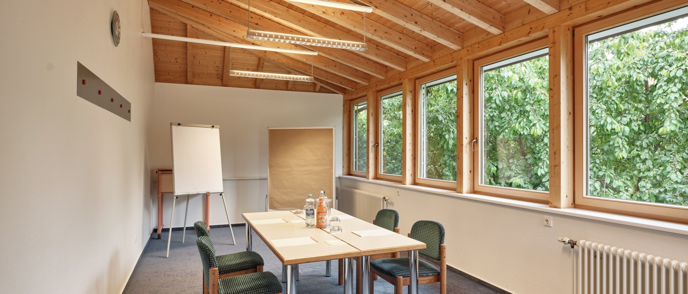 Ein heller Seminarraum mit Holzdach, Tisch und Stühlen. Große Fenster bieten Blick ins Grüne. Ein Flipchart steht bereit., © Badhotel Restaurant Stauferland
