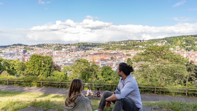 Zwei Personen sitzen auf einer Wiese und blicken auf die Stadt Stuttgart. Im Hintergrund sind Hügel und ein blauer Himmel mit Wolken zu sehen., © © Stuttgart-Marketing GmbH, Martina Denker Zwei Personen sitzen auf einer Wiese und blicken auf die Stadt Stuttgart. Im Hintergrund sind Hügel und ein blauer Himmel mit Wolken zu sehen., © © Stuttgart-Marketing GmbH, Martina Denker