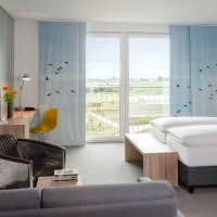 Modernes Hotelzimmer mit gro&szlig;em Bett, Schreibtisch, Sessel und Balkon mit Aussicht auf Landschaft. Dekor mit blauen Vorh&auml;ngen und Vogelmotiven., &copy; Essential by Dorint