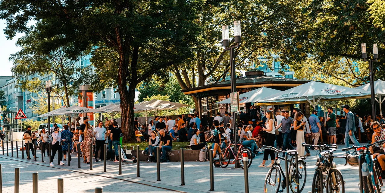 Lebendiger Platz vor dem Palast der Republik in Stuttgart mit vielen Menschen, Fahrrädern und Sonnenschirmen im Schatten der Bäume., © SMG, Sarah Schmid Lebendiger Platz vor dem Palast der Republik in Stuttgart mit vielen Menschen, Fahrrädern und Sonnenschirmen im Schatten der Bäume., © SMG, Sarah Schmid