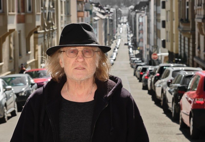 Ein Mann mit Hut und Brille steht auf einer belebten Straße mit geparkten Autos und Gebäuden im Hintergrund., © Rosenau Kultur e.V. Ein Mann mit Hut und Brille steht auf einer belebten Straße mit geparkten Autos und Gebäuden im Hintergrund., © Rosenau Kultur e.V.