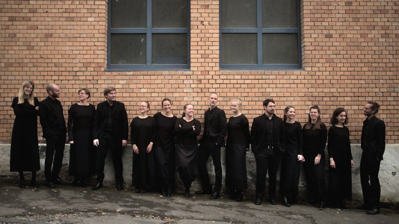 Eine Gruppe von 14 Personen in schwarzer Kleidung steht lächelnd vor einer Backsteinmauer mit Fenstern., © Kammerchor Vokalkunst / Daniel Radde Eine Gruppe von 14 Personen in schwarzer Kleidung steht lächelnd vor einer Backsteinmauer mit Fenstern., © Kammerchor Vokalkunst / Daniel Radde