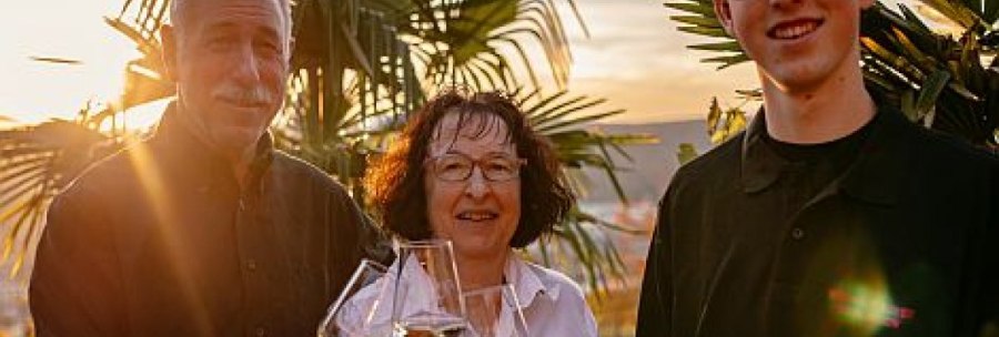 Three people are clinking wine glasses, surrounded by palm trees at sunset., © Weingut Bayer Three people are clinking wine glasses, surrounded by palm trees at sunset., © Weingut Bayer