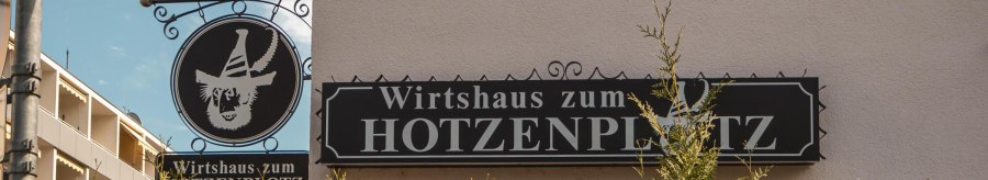 Schild des Wirtshauses zum Hotzenplotz an der Lindensp&uuml;rstra&szlig;e, umgeben von gr&uuml;nen B&uuml;schen und einem blauen Himmel im Hintergrund., &copy; SMG. Sarah Schmid