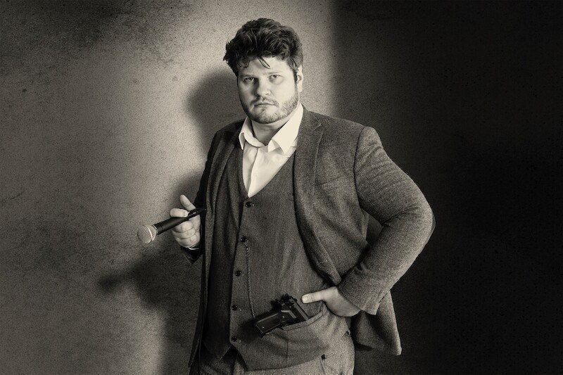 Ein Mann in einem Anzug hält ein Mikrofon und eine Kamera. Er steht vor einem neutralen Hintergrund und schaut ernst in die Kamera., © Rosenau Kultur e.V. Ein Mann in einem Anzug hält ein Mikrofon und eine Kamera. Er steht vor einem neutralen Hintergrund und schaut ernst in die Kamera., © Rosenau Kultur e.V.