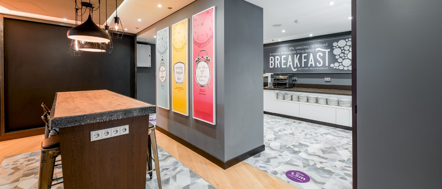 Moderner Frühstücksbereich mit Holztisch, Hockern und dekorativen Postern. Im Hintergrund ein Buffetbereich mit Geschirr und Frühstücksangebot., © Premier Inn Moderner Frühstücksbereich mit Holztisch, Hockern und dekorativen Postern. Im Hintergrund ein Buffetbereich mit Geschirr und Frühstücksangebot., © Premier Inn