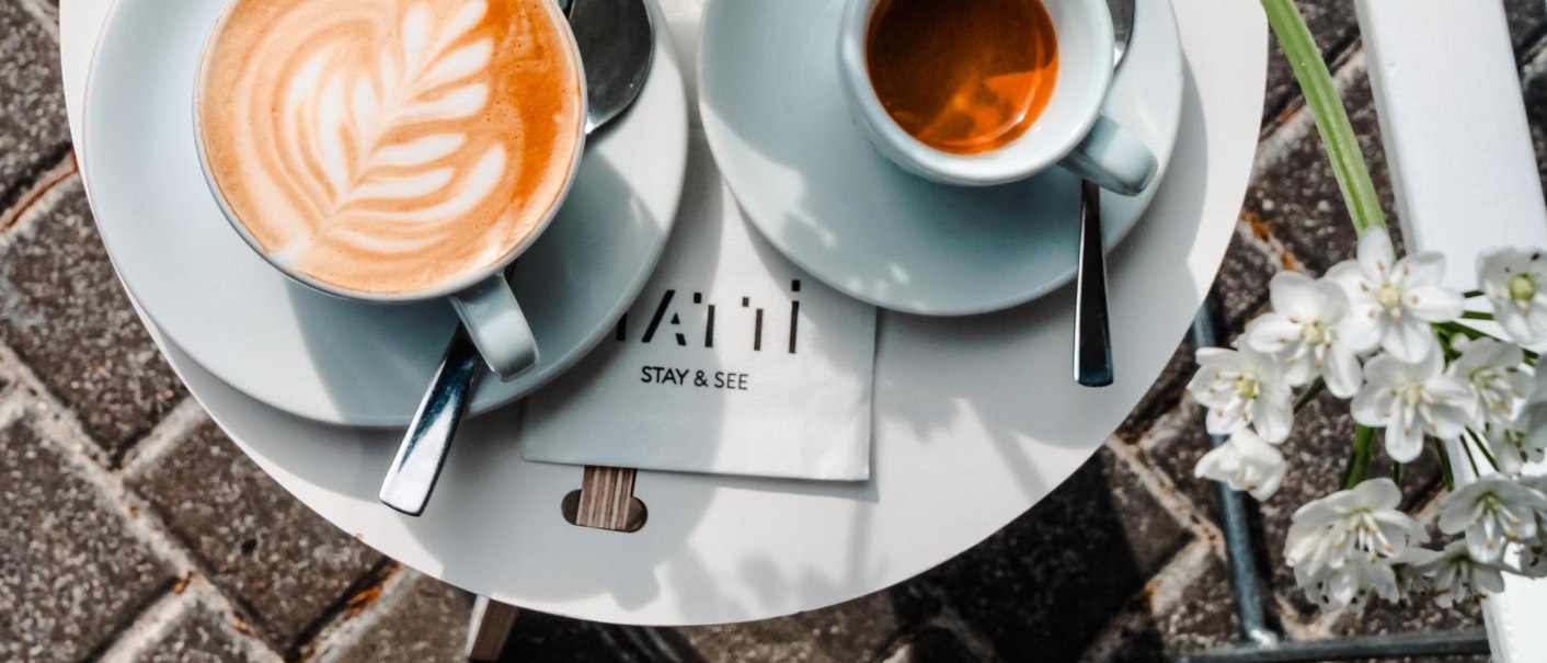 Ein runder Tisch mit Cappuccino und Espresso in Tassen, daneben weiße Blumen und eine Serviette mit der Aufschrift "STAY & SEE"., © Tatti Stay & See