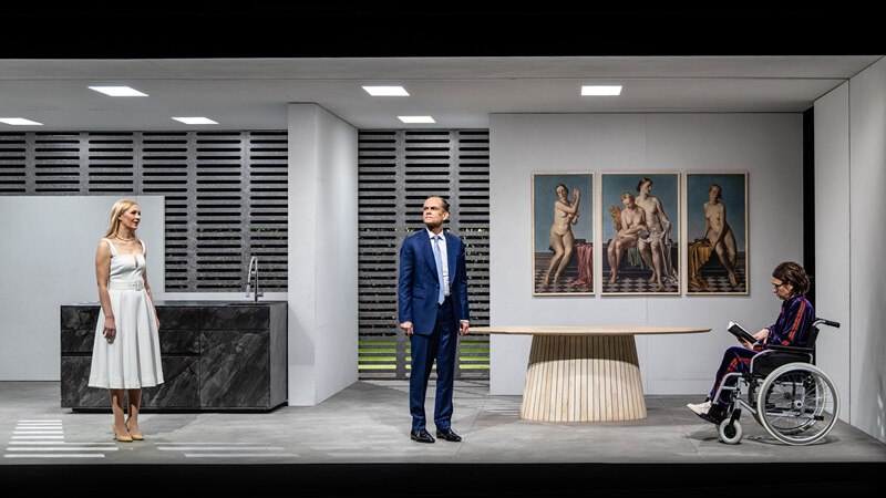 Theater scene in a modern room with three people. A woman in a wheelchair reads while a man and a woman stand. Works of art on the wall., &copy; W&uuml;rttembergische Staatstheater Stuttgart