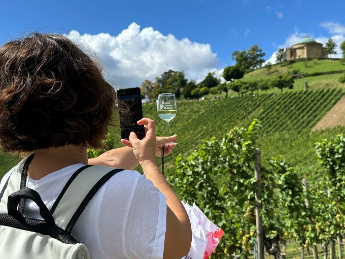 Eine Frau fotografiert ein Weinglas vor einem malerischen Weinberg. Im Hintergrund ist eine Kapelle auf einem Hügel zu sehen., © Weingenuss Stuttgart Eine Frau fotografiert ein Weinglas vor einem malerischen Weinberg. Im Hintergrund ist eine Kapelle auf einem Hügel zu sehen., © Weingenuss Stuttgart