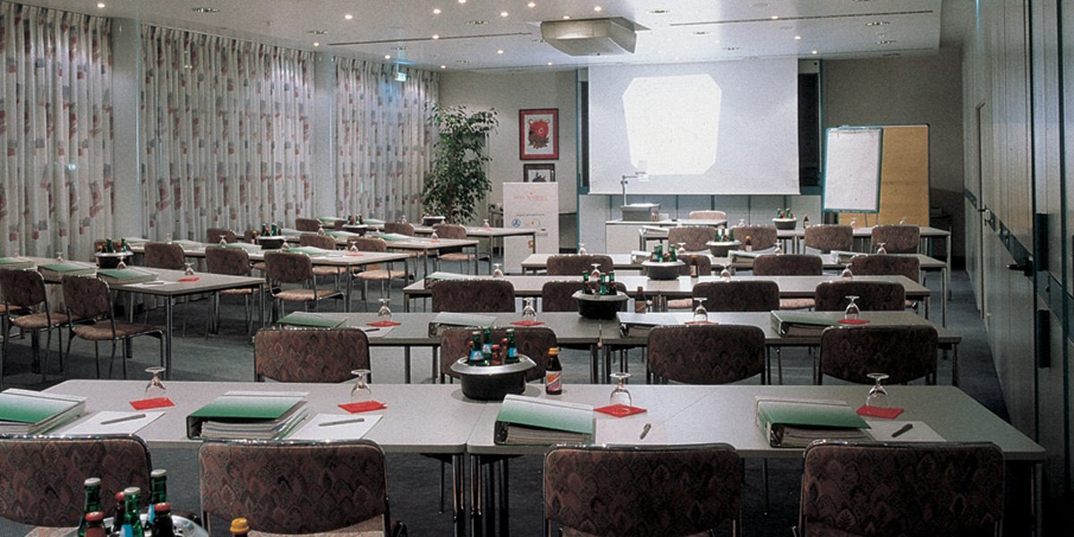 An empty conference room with tables, chairs, a projector and a flipchart. There are folders and bottles of water on the tables., © © H-Hotels.com An empty conference room with tables, chairs, a projector and a flipchart. There are folders and bottles of water on the tables., © © H-Hotels.com