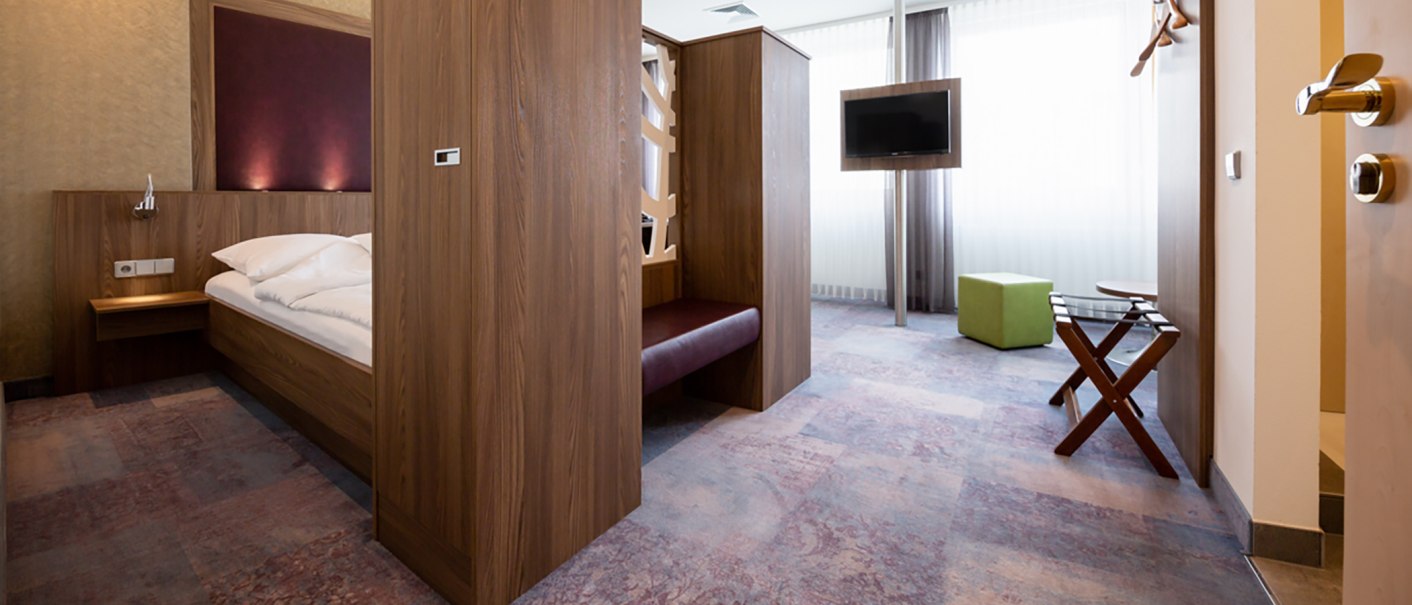 Modernes Hotelzimmer mit Holzmöbeln, Bett, Fernseher und grünem Hocker. Helle Vorhänge und Teppichboden ergänzen das Design., © Kronenhotel Stuttgart