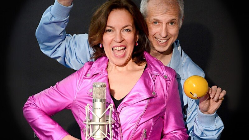Zwei Personen posieren fröhlich mit Maracas und einem Mikrofon vor schwarzem Hintergrund. Eine Person trägt eine pinke Jacke, die andere eine blaue., © Theaterhaus Stuttgart e.V.