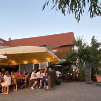 Menschen sitzen unter großen Sonnenschirmen in einem gemütlichen Hof mit roten Dächern und Bäumen., © Weinhof Zaiß