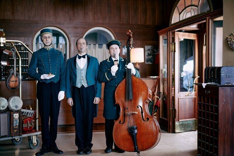 Drei Männer in Hoteluniformen stehen in einer Lobby. Einer hält einen Kontrabass, die anderen tragen Tabletts. Die Szene wirkt wie aus einem alten Hotel., © Theaterhaus Stuttgart e.V.