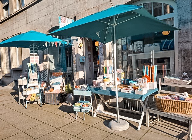 Ein Laden im Stuttgarter Süden mit blauen Sonnenschirmen. Stoffartikel und Dekorationen sind auf Tischen und Stühlen im Freien ausgestellt., © Cosima Chiton, Stuttgart