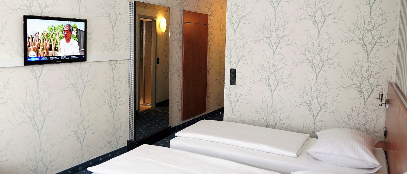 A hotel room with two single beds, a TV mounted on the wall, and a large mirror. The walls are covered with wallpaper featuring a tree-like pattern., &copy; TOMAS