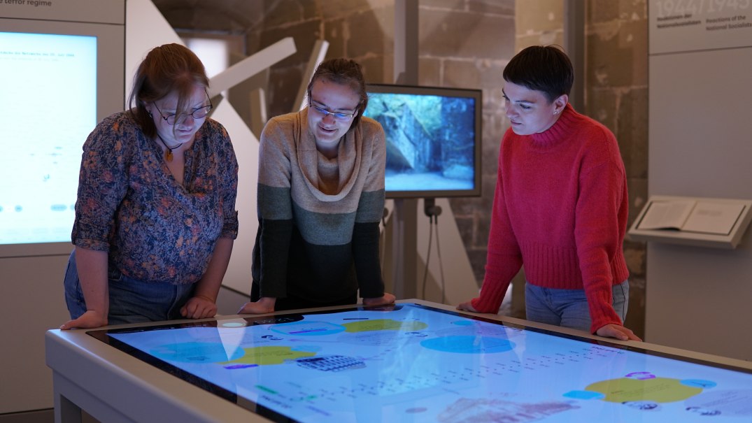 Drei Personen stehen um einen interaktiven Medientisch in einer Ausstellung und betrachten die darauf angezeigten Informationen., © HdGBW Drei Personen stehen um einen interaktiven Medientisch in einer Ausstellung und betrachten die darauf angezeigten Informationen., © HdGBW