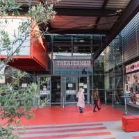 Entrance to the Theaterhaus Stuttgart with red floor, glass facade and posters. Two people enter the building. Plants and modern architecture are visible., © Stuttgart-Marketing GmbH, wpsteinheisser