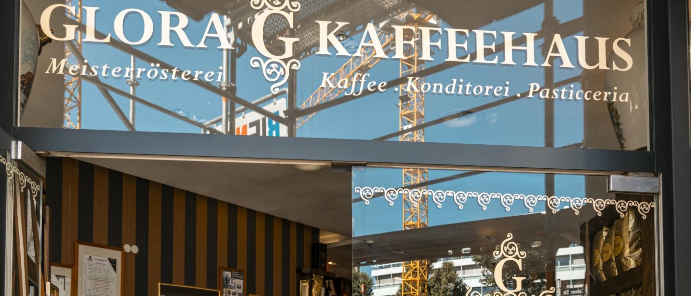 Eingang des Glora Kaffeehauses mit dekorativem Schriftzug auf der Glasfront. Im Hintergrund sind Baukräne zu sehen., © SMG, Sarah Schmid Eingang des Glora Kaffeehauses mit dekorativem Schriftzug auf der Glasfront. Im Hintergrund sind Baukräne zu sehen., © SMG, Sarah Schmid