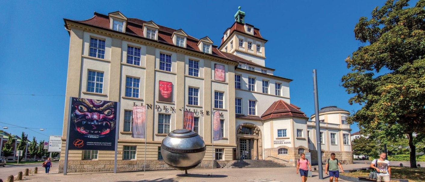 The Linden Museum in Stuttgart in sunny weather. Posters and a large silver sphere can be seen in front of the building. People stroll past., © © Stuttgart-Marketing GmbH