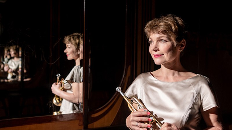 Eine Frau in einem eleganten, hellen Kleid h&auml;lt eine Trompete und steht vor einem Spiegel, der ihr Bild mehrfach reflektiert., &copy; Altes Schauspielhaus und Kom&ouml;die im Marquardt e.V.