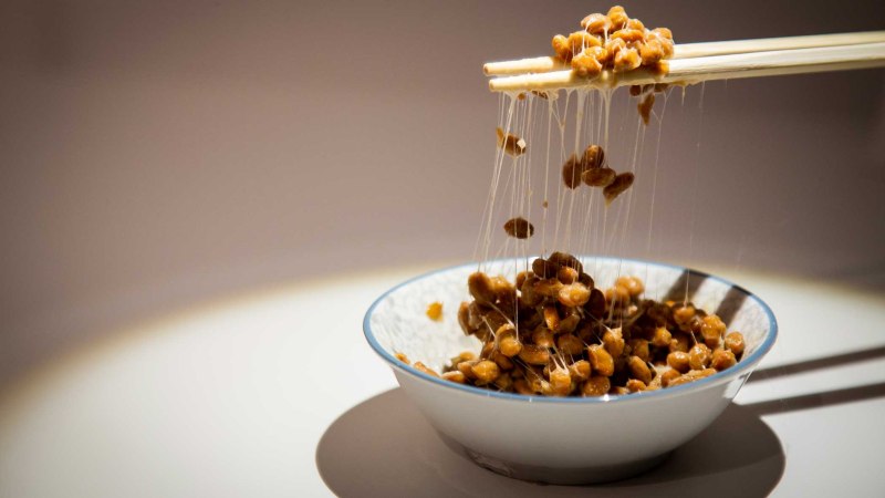 Fermentierte Sojabohnen (Natto) werden mit Stäbchen aus einer Schale gehoben, Fäden ziehen sich zwischen den Bohnen., © Foto: Anja Barte Telin