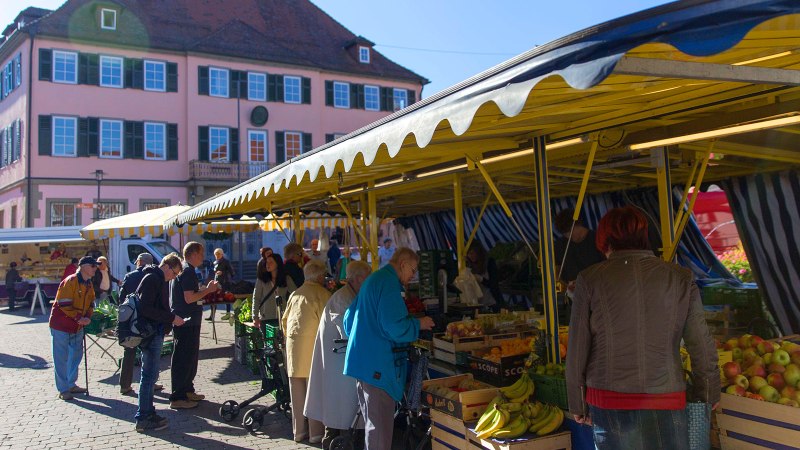 People shop at a market in Murrhardt. Yellow market stalls offer fruit and vegetables. A large pink building in the background., © Stuttgart-Marketing GmbH, Achim Mende People shop at a market in Murrhardt. Yellow market stalls offer fruit and vegetables. A large pink building in the background., © Stuttgart-Marketing GmbH, Achim Mende