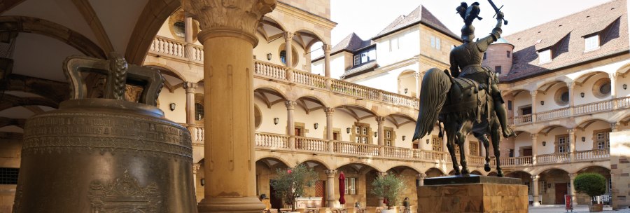 Innenhof eines alten Schlosses mit Reiterstatue, großer Glocke und Arkaden. Tische und Stühle stehen im Hof, umgeben von historischen Gebäuden., © Stuttgart-Marketing GmbH, Christoph Düpper