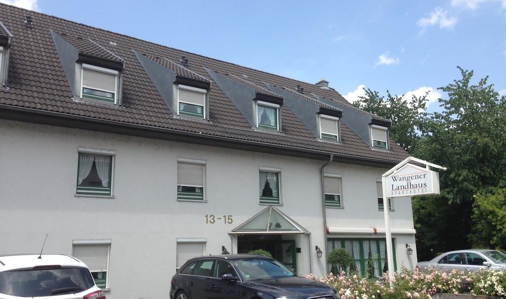 Das Aparthotel Wangener Landhaus mit grauem Dach und weißen Wänden. Vor dem Gebäude sind Autos geparkt. Ein Schild zeigt den Hotelnamen., © Aparthotel Wangener Landhaus Das Aparthotel Wangener Landhaus mit grauem Dach und weißen Wänden. Vor dem Gebäude sind Autos geparkt. Ein Schild zeigt den Hotelnamen., © Aparthotel Wangener Landhaus