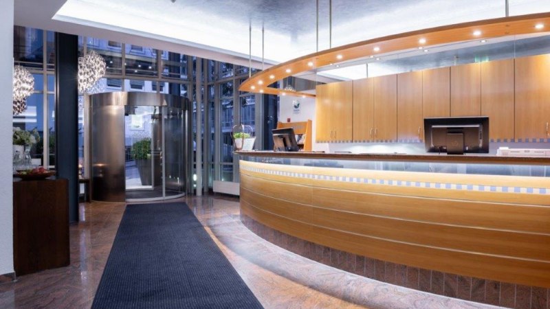 Modern hotel reception with curved wooden counter, illuminated reception area and revolving door in the background. Elegant, inviting ambience., &copy; Kronen Hotel GmbH