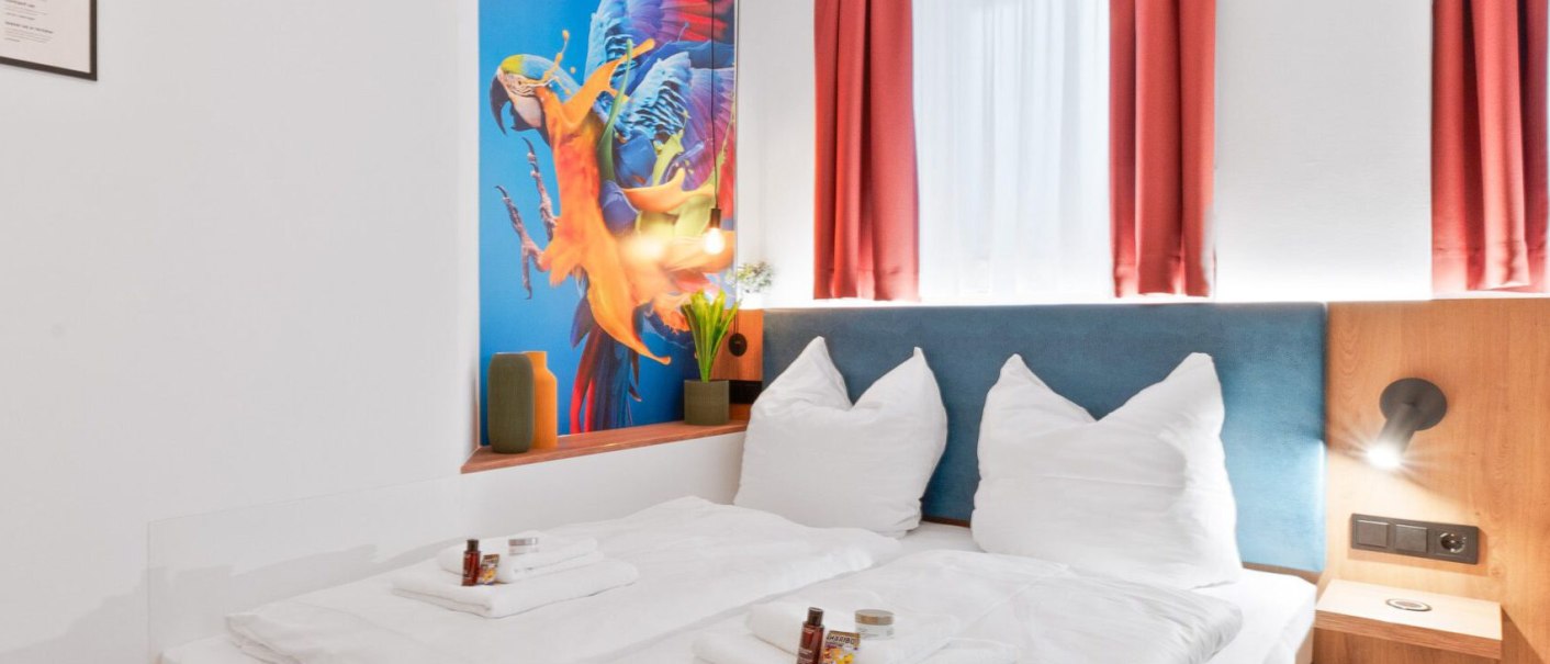Modernes Doppelzimmer mit weißem Bett, Papageien-Wandbild, roten Vorhängen und gelbem Hocker. Pflegeprodukte auf dem Bett., © JUNGLE N°5 GmbH Modernes Doppelzimmer mit weißem Bett, Papageien-Wandbild, roten Vorhängen und gelbem Hocker. Pflegeprodukte auf dem Bett., © JUNGLE N°5 GmbH