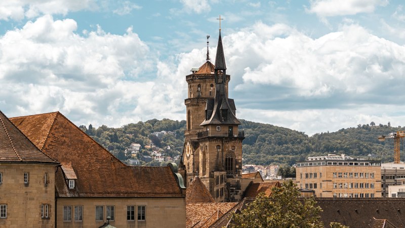 Die Stiftskirche mit ihrem markanten Turm erhebt sich über die Dächer der Stadt, umgeben von grünen Hügeln und einem bewölkten Himmel., © Stuttgart-Marketing GmbH, Sarah Schmid Die Stiftskirche mit ihrem markanten Turm erhebt sich über die Dächer der Stadt, umgeben von grünen Hügeln und einem bewölkten Himmel., © Stuttgart-Marketing GmbH, Sarah Schmid