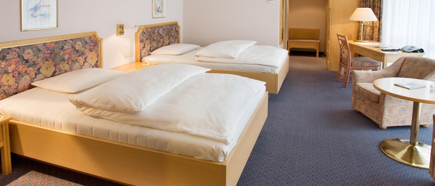 A spacious hotel room with two single beds, floral headboard, desk, armchair and table. Light wood furniture and carpeted floor., &copy; Erikson Hotel