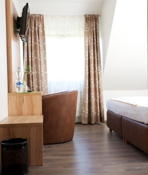 A cozy hotel room with a double bed, brown armchair, TV on the wall, curtains and wooden furniture., © Hotel Reinhardtshof Garni A cozy hotel room with a double bed, brown armchair, TV on the wall, curtains and wooden furniture., © Hotel Reinhardtshof Garni