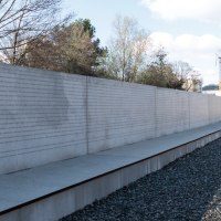 Eine lange Betonwand mit eingravierten Namen steht neben stillgelegten Bahngleisen unter einem bew&ouml;lkten Himmel., &copy; Foto-Kunst Andreas Keller
