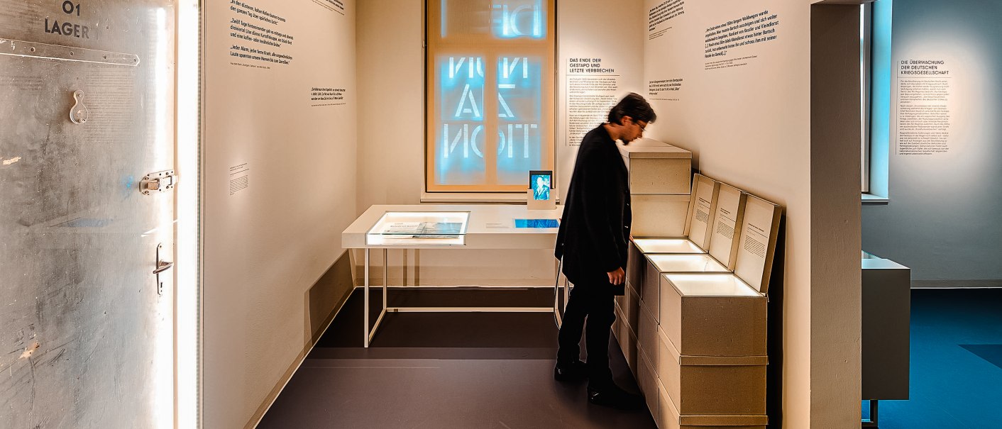 Ein Mann in einem Ausstellungsraum des HdG-Hotel Silber, betrachtet beleuchtete Vitrinen. An den Wänden sind informative Texte angebracht., © Daniel Stauch